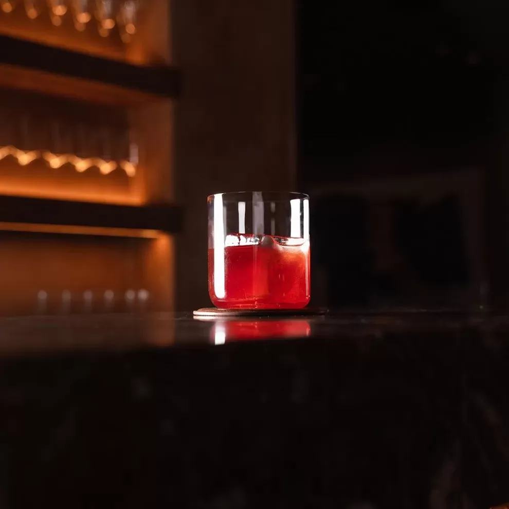 A glass of red cocktail with ice sits on a dark tabletop. The dimly lit background features shelves with glasses, giving a cosy and intimate atmosphere. Served at Akoko, Fitzrovia.