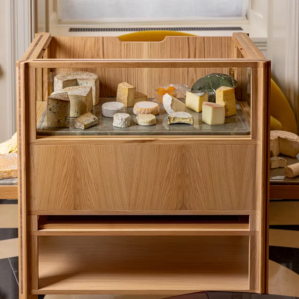 A wooden cheese cart displaying a variety of cheeses in different shapes, colors, and textures. The cart is positioned indoors, with natural light highlighting the assortment of cheeses at Mauro Colagreco at Raffles London at The OWO, Whitehall.