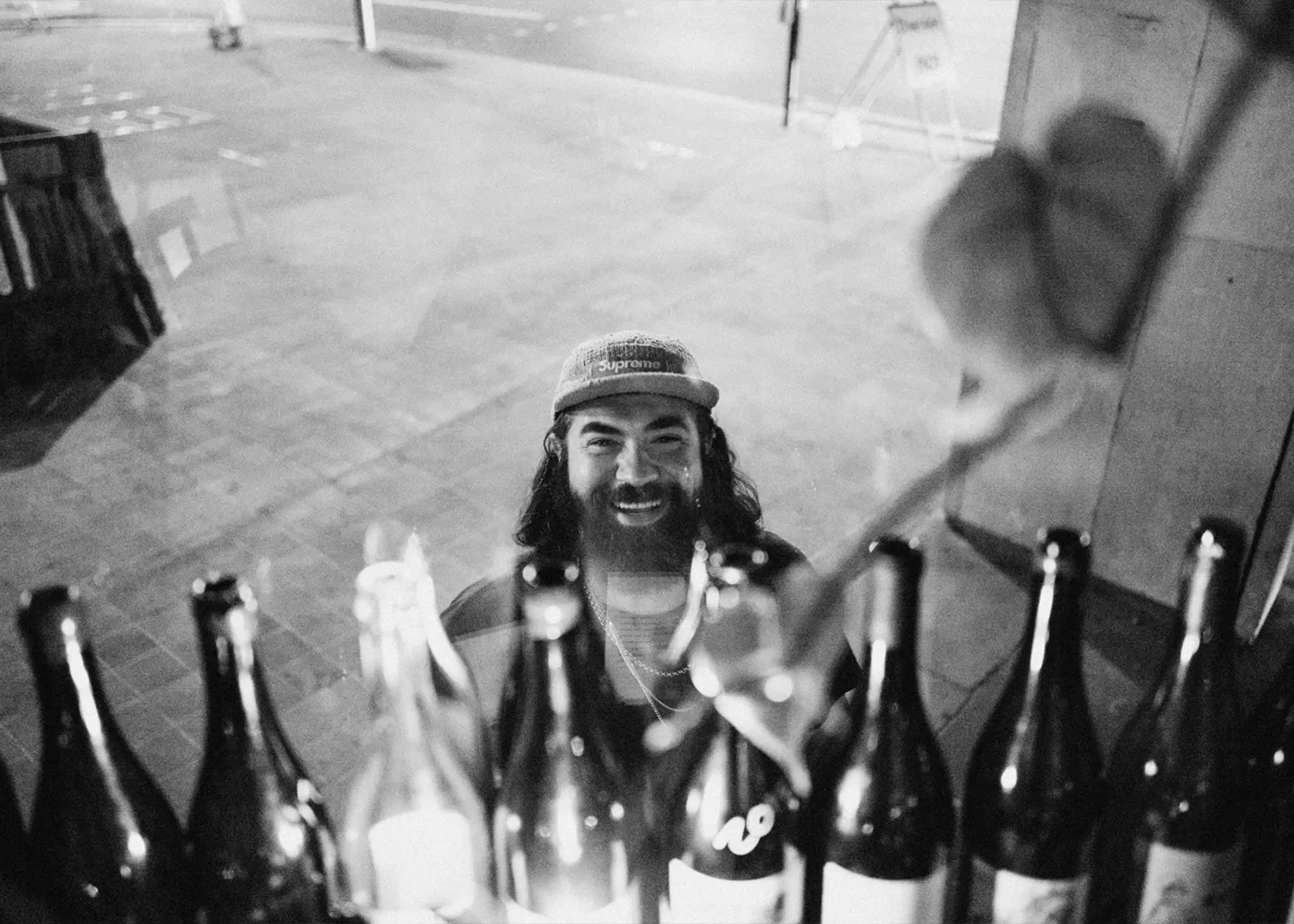 A bearded man in a cap smiles behind a row of empty glass bottles on a window sill, with a blurred street scene visible in the background. The photo is in black and white.