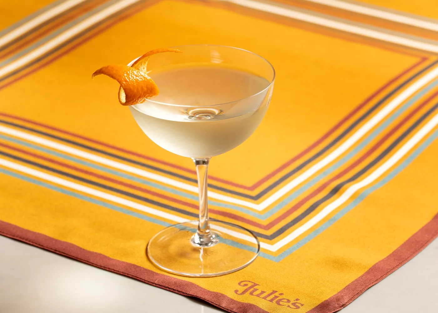 A clear cocktail in a stemmed glass garnished with a twist of orange peel sits on a yellow, striped napkin with the name Julies embroidered in the corner.
