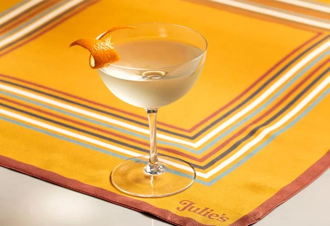 A clear cocktail in a coupe glass garnished with an orange twist sits on a yellow, striped placemat with the name Julies embroidered in the corner.
