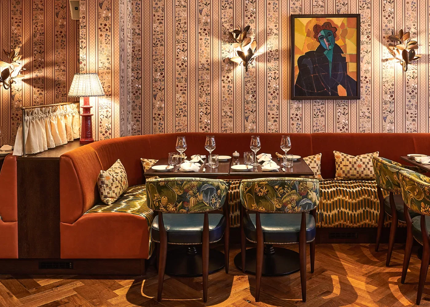 Cosy restaurant interior featuring a curved orange booth with patterned pillows, surrounded by green, floral-upholstered chairs, a patterned wallpaper, and a colorful abstract painting. Tables are set with glasses and napkins.