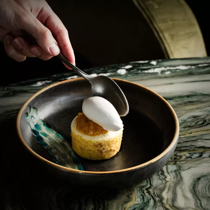 A hand holds a spoon above a plated dessert featuring a small cake topped with a dollop of fruit compote and a quenelle of white cream or sorbet, served on a ceramic dish with a green marble table underneath.