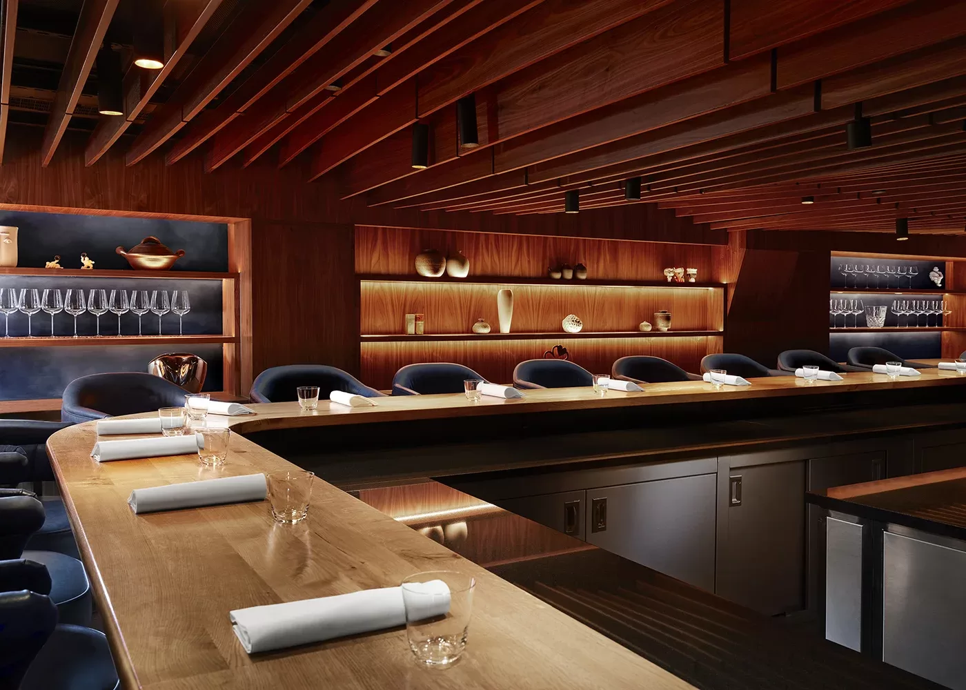The modern, elegant restaurant interior of Kitchen Table, Fitzrovia, with a long, wooden counter surrounded by navy blue chairs. Soft, warm lighting highlights glassware and decorative items on wooden shelves, creating a cozy, sophisticated ambiance.
