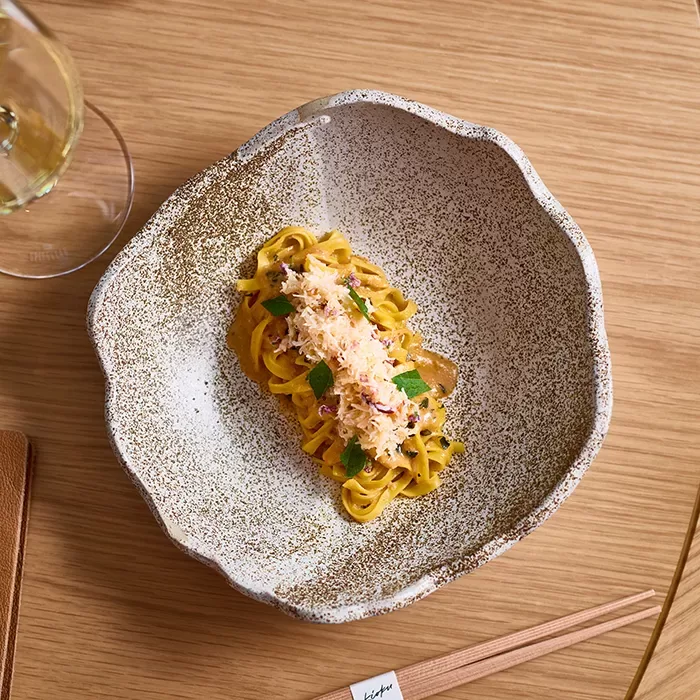 Spaghetti topped with crab served in a scalloped edged bowl on a wooden table with a glass of white wine at restaurant Kioku by Endo in London.
