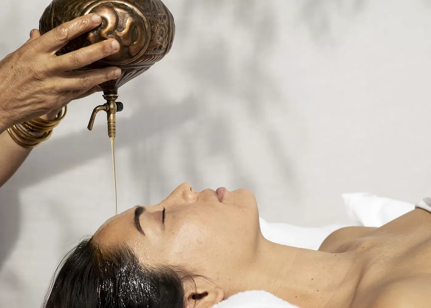 A person lies on their back with eyes closed as oil is gently poured onto their forehead from a brass vessel, suggesting a relaxing spa or Ayurvedic treatment.