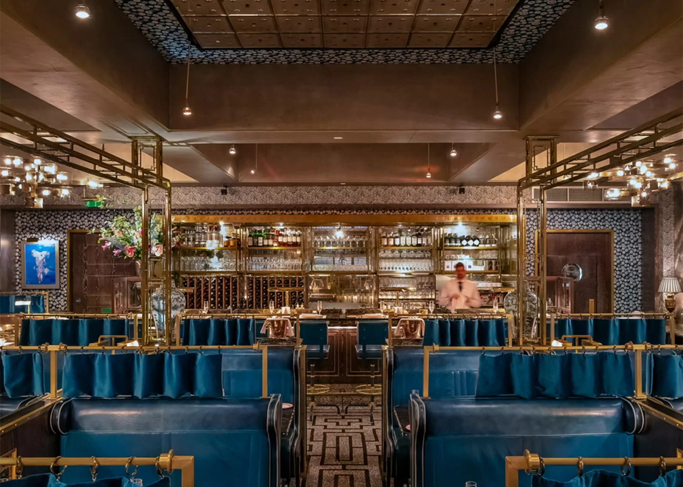 Luxurious restaurant interior with blue velvet seating, gold accent decor, and a patterned floor. A bartender stands behind a well-stocked, illuminated bar with glass shelves in the background at Bob Bob Ricard Soho London, London.
