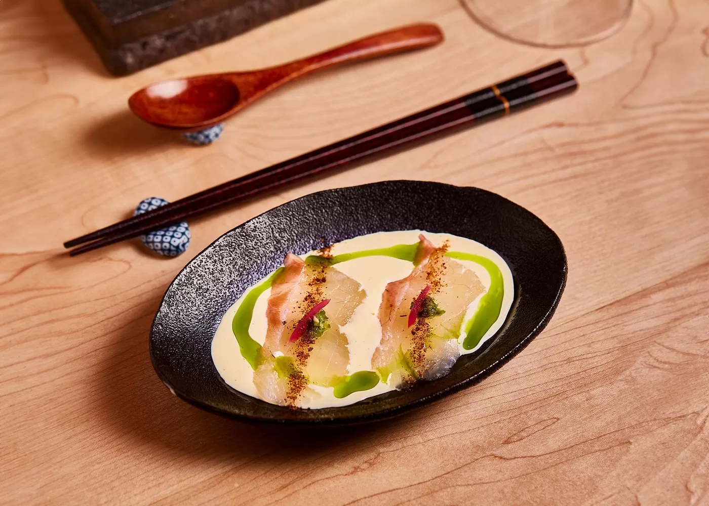 A beautifully plated gourmet dish featuring thin slices of fish, drizzled with vibrant green sauce and a creamy base, elegantly presented in a black dish. A wooden spoon and chopsticks rest on the side, adding a refined touch to the presentation served at Juno Omakase, London.