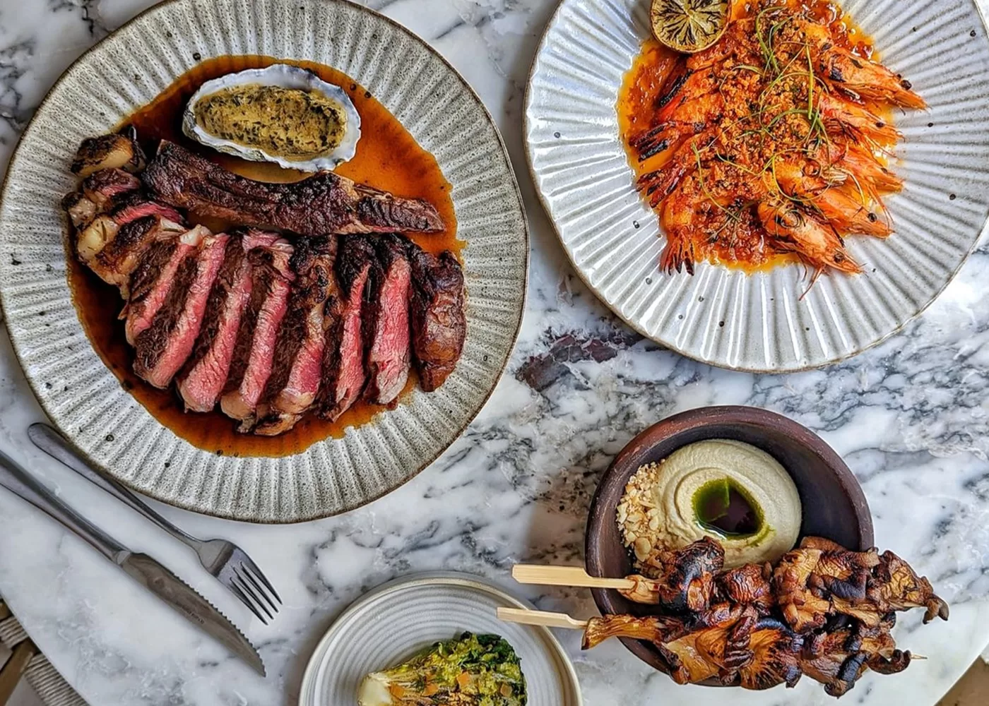 A marble table with plates of food: sliced steak with sauce, grilled prawns with lemon garnish, skewered grilled chicken with hummus, and a small bowl of greens. Silverware is placed beside the plates at Milk Beach, London.