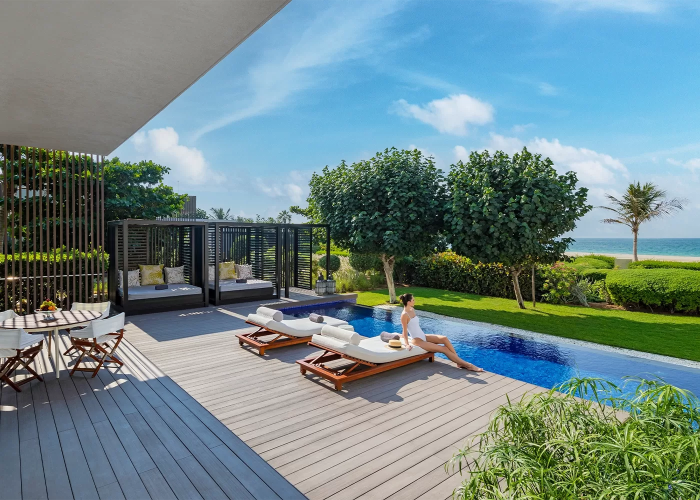 A woman relaxes on a lounge chair by an outdoor pool overlooking the ocean. The scene includes lush greenery, modern cabanas, and a small dining table on a wooden deck under a clear blue sky at The Oberoi Beach Resort, Al Zorah, Dubai.