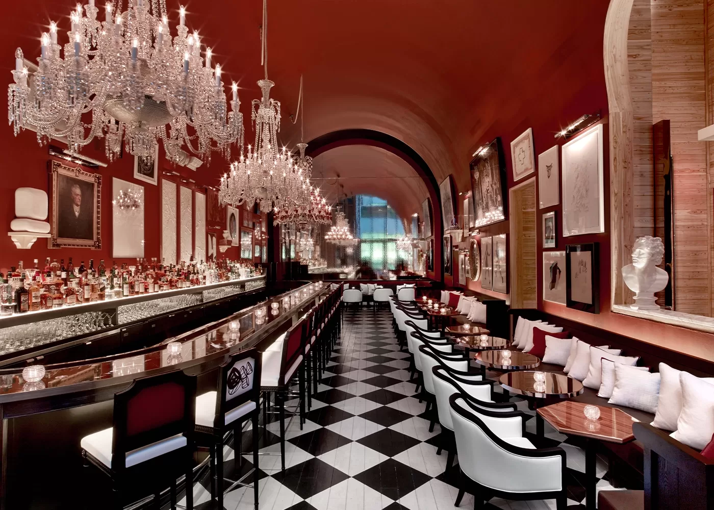 The image shows an elegant, dimly lit bar with long black countertops and plush white stools. Crystal chandeliers hang from a high arched ceiling, and the floor is a black-and-white checkered pattern. Artwork decorates the deep red walls.