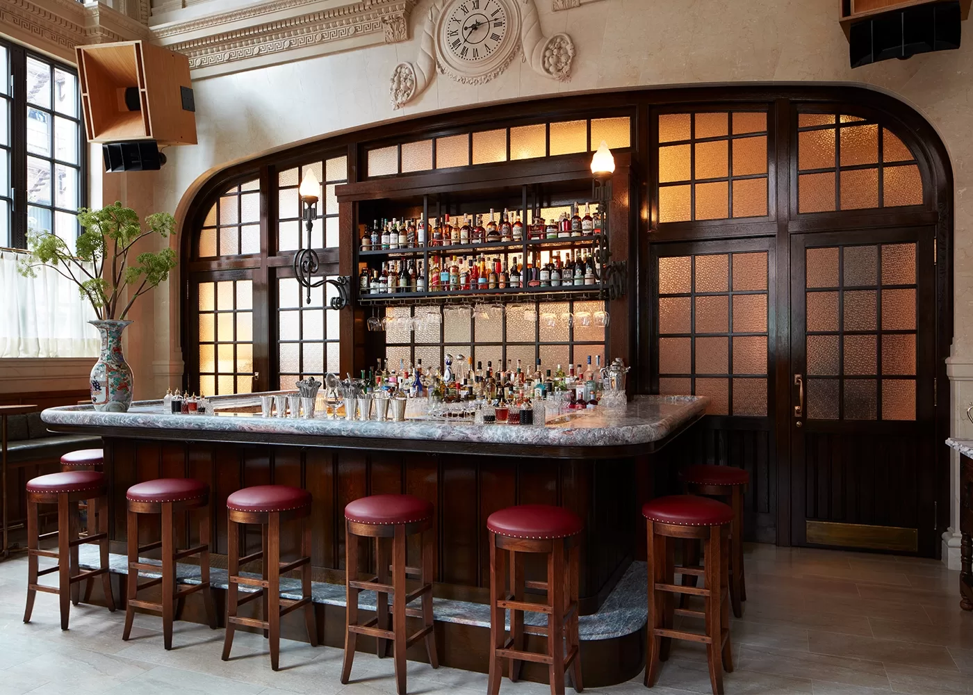 A stylish bar interior featuring a marble-topped bar with red cushioned stools. The back wall showcases shelves filled with various liquor bottles. Large windows and decorative elements create a warm, inviting atmosphere.