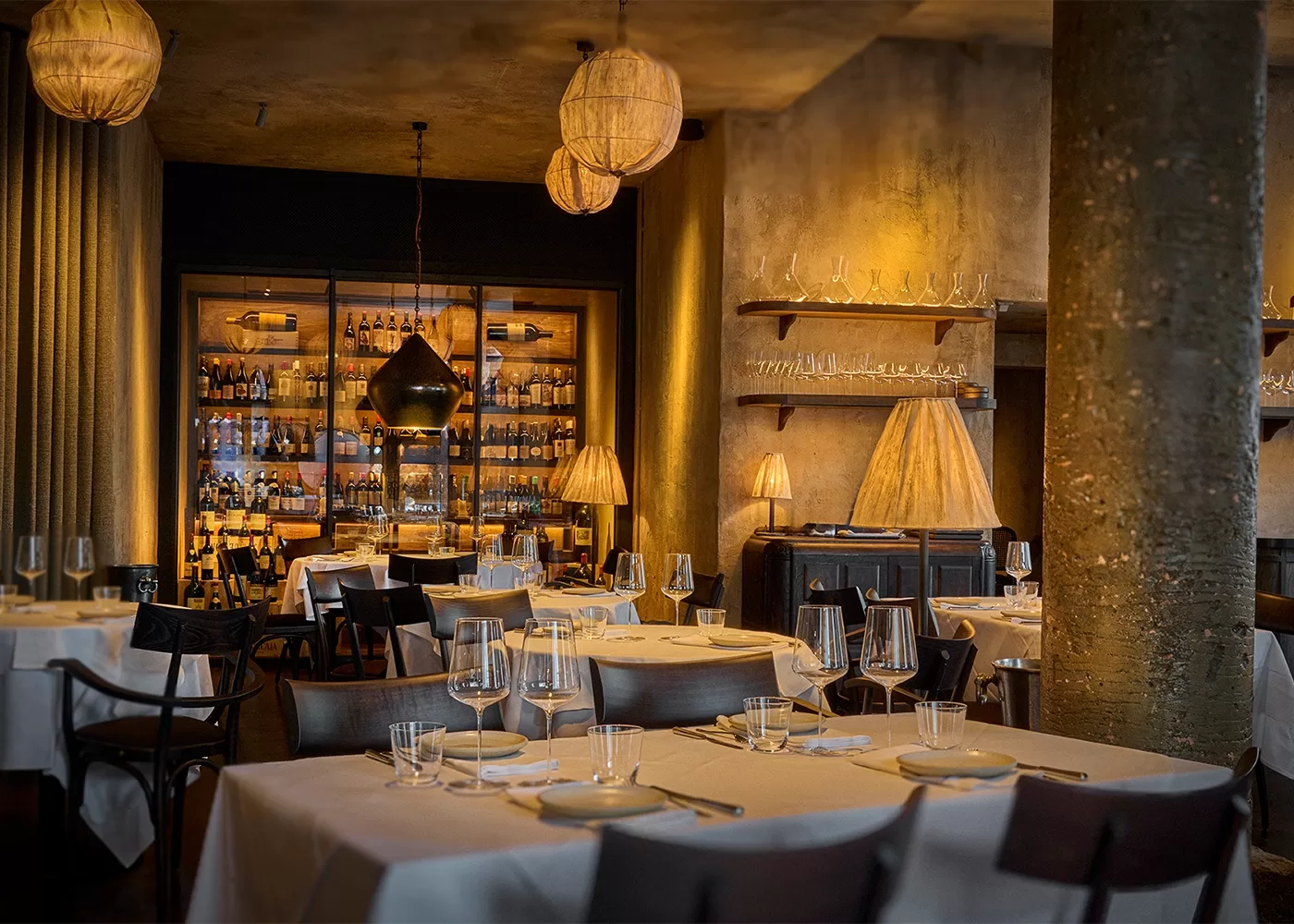 A dimly lit, elegant restaurant with white tablecloths, glassware, and dark chairs. Warm lighting from unique pendant lamps and a wall of shelves holding bottles creates an inviting ambiance at Fantômas, London.