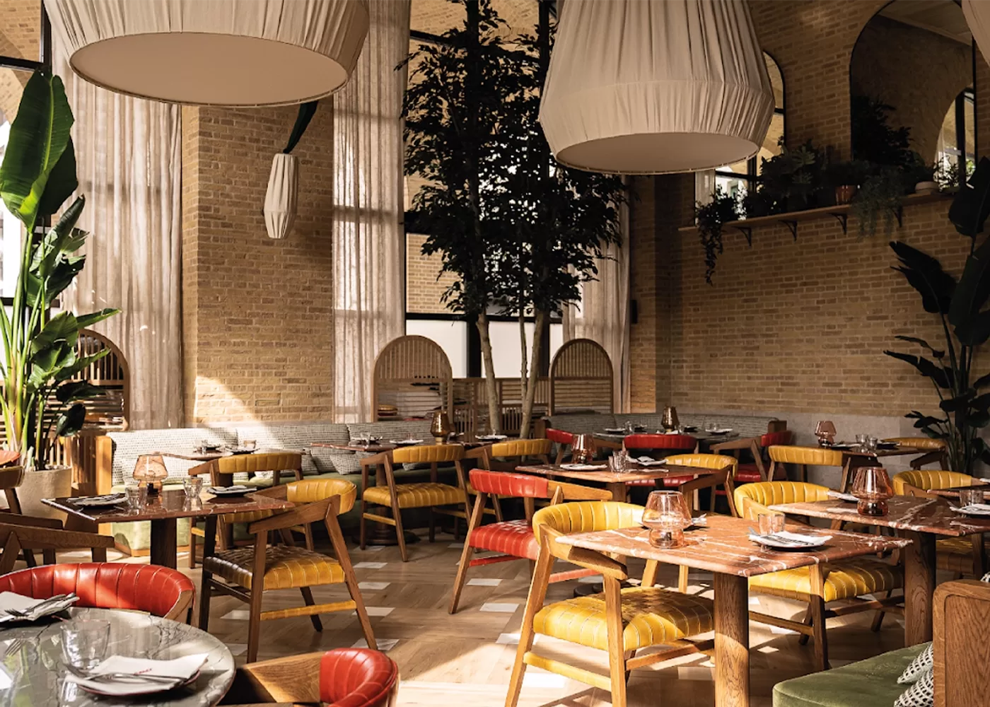A cosy restaurant interior with rustic brick walls and large windows. There are wooden tables and colorful cushioned chairs, surrounded by plants. Large pendant lights hang from the high ceiling, creating a warm and inviting atmosphere at The Campaner, London.