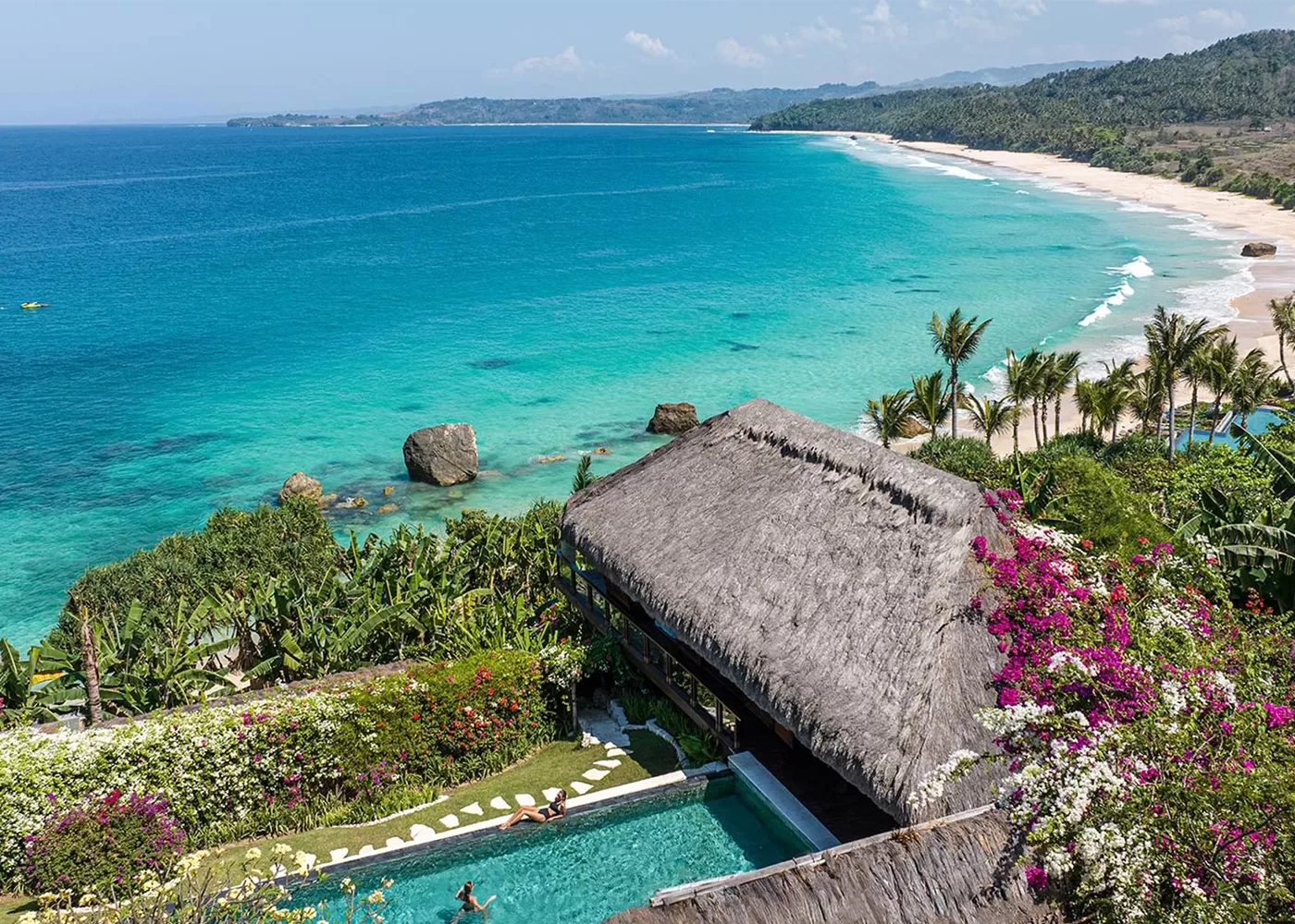 A stunning coastal view of a turquoise ocean with white sandy beaches. In the foreground, a tropical villa with a thatched roof overlooks a pool surrounded by vibrant flowers and lush greenery.