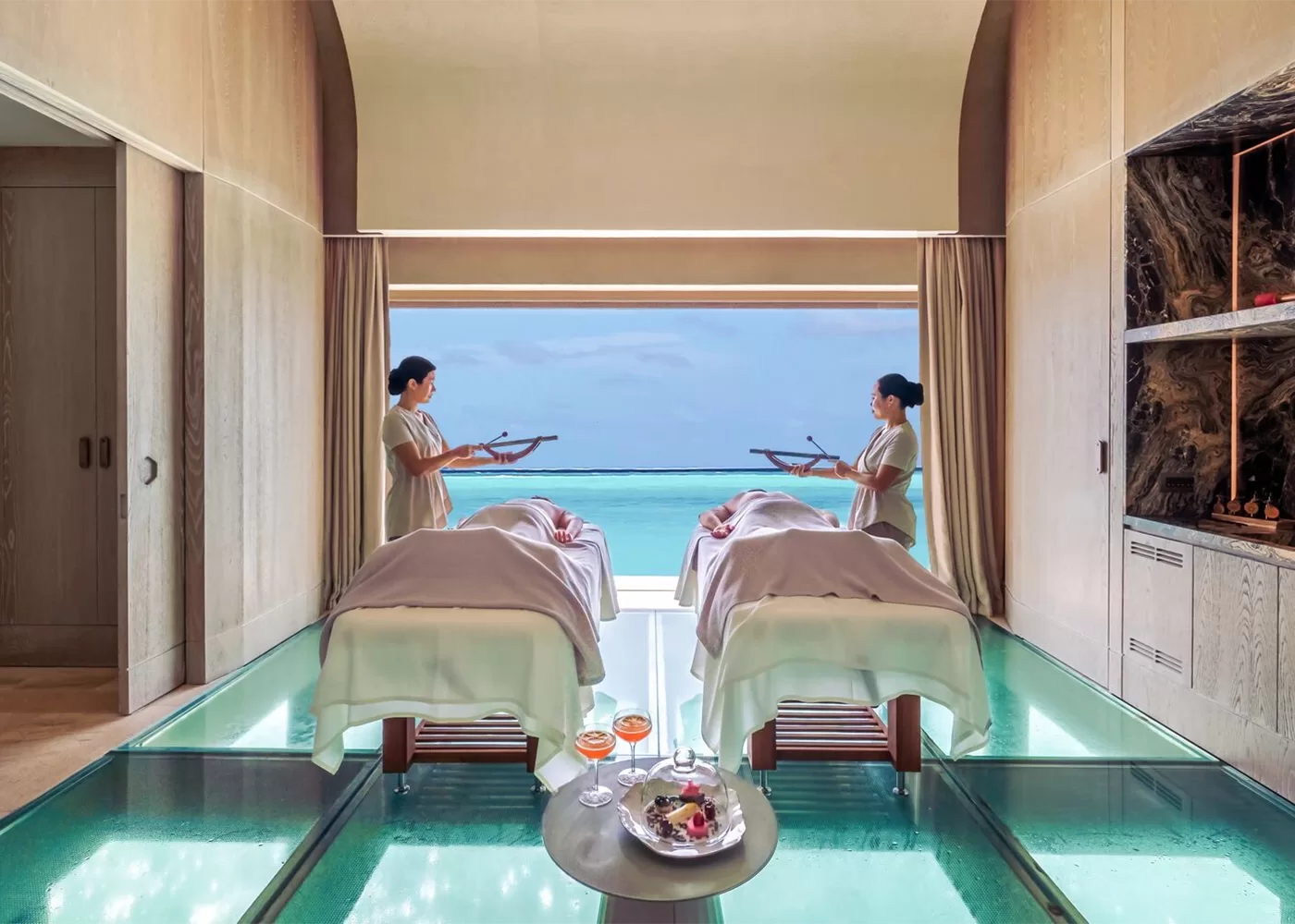 Two massage tables are set in a luxurious spa room with a glass floor overlooking a turquoise ocean view. Two therapists stand holding trays beside the tables. The ambiance is serene and relaxing, with soft lighting and wooden decor.