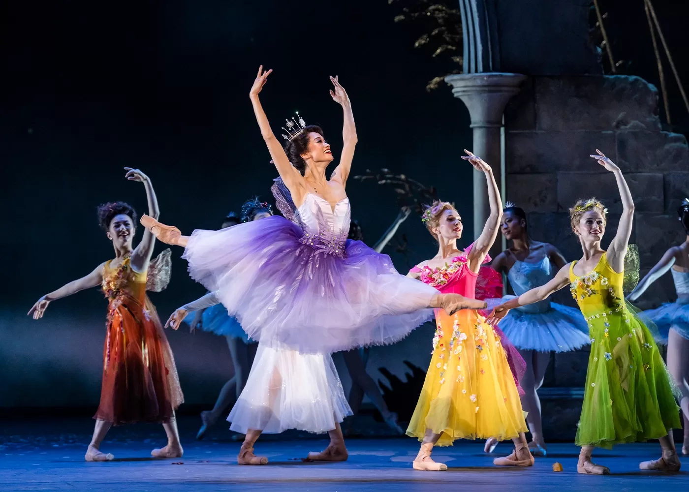 A ballerina in a purple and white tutu stands on stage, surrounded by dancers in tutus. They hold sparkling wands, poised elegantly on a staircase backdrop, creating a magical, fairytale-like scene at Royal Ballet & Opera’s Cinderella.