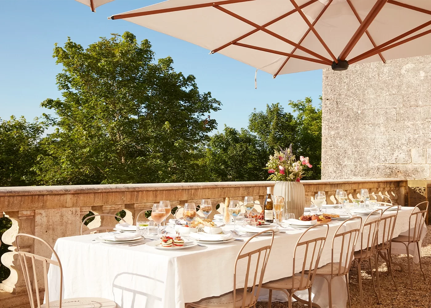 A beautifully set outdoor dining table with a white tablecloth and various dishes is under a large umbrella. Elegant chairs surround the table. In the background, there are lush green trees and a stone wall, suggesting a scenic, sunny setting.