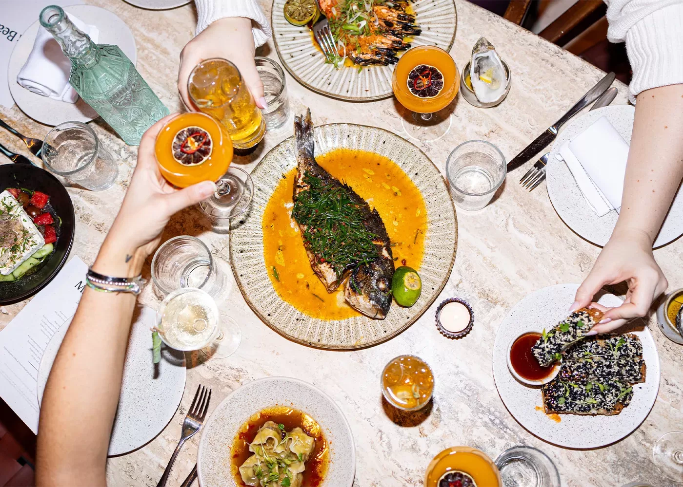 A top view of a dining table with a variety of dishes, including a whole cooked fish, grilled prawns, appetizers, and drinks. Three people are reaching for food and holding glasses, creating a lively and social dining atmosphere at Milk Beach Soho Soho London, London.