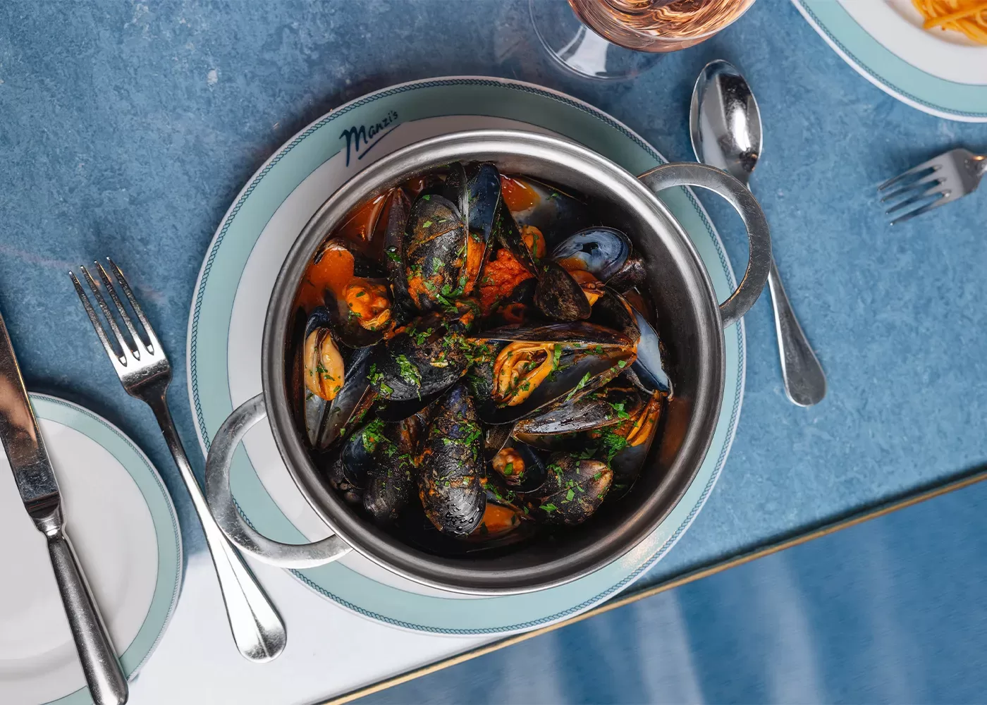 A pot of steamed mussels in a rich broth garnished with herbs, set on a blue table. Silverware is arranged neatly around the dish, and a glass of rosé wine is visible in the background  at Manzi’s, Soho, London.