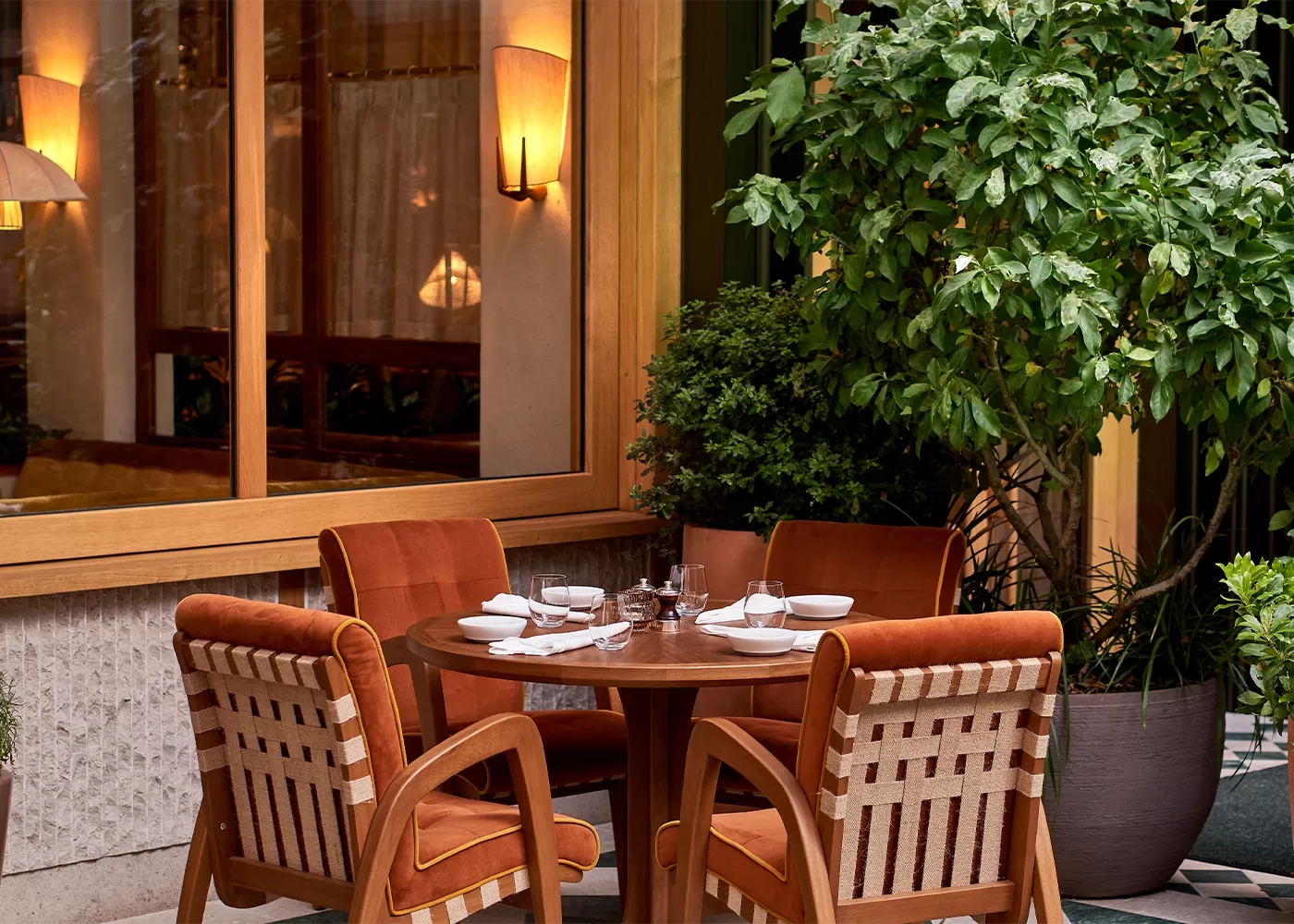 A cosy outdoor dining area features a round wooden table set for four, with burnt orange cushioned chairs. The setting includes white tableware and is surrounded by green plants, near a window with warm lighting at Dovetale, London.