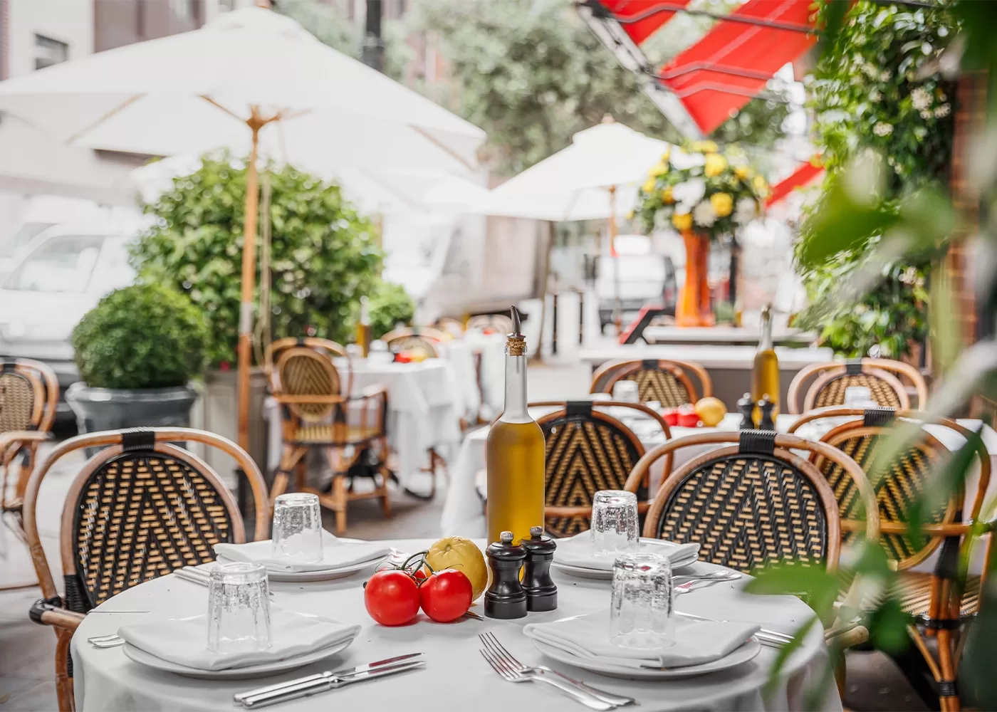 An outdoor restaurant patio with tables set with white tablecloths, plates, cutlery, olive oil, and seasonings. Rattan chairs surround the tables, and red umbrellas provide shade. Tomatoes are placed on one table as decoration at LPM, London.
