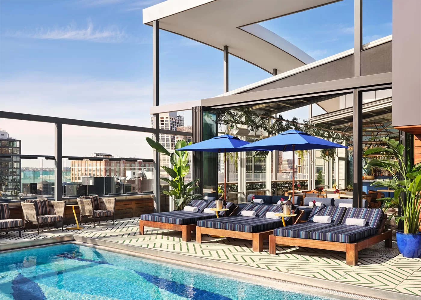 Blue skies overlooking a swimming pool with a glass wall around it overlooking buildings. Blue striped sun loungers with blue parasols on the side.