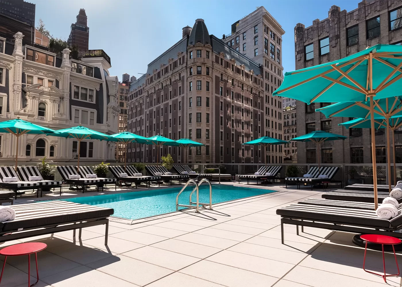 Glimmering blue pool surrounded by New York buildings and turquoise parasols over sun loungers,