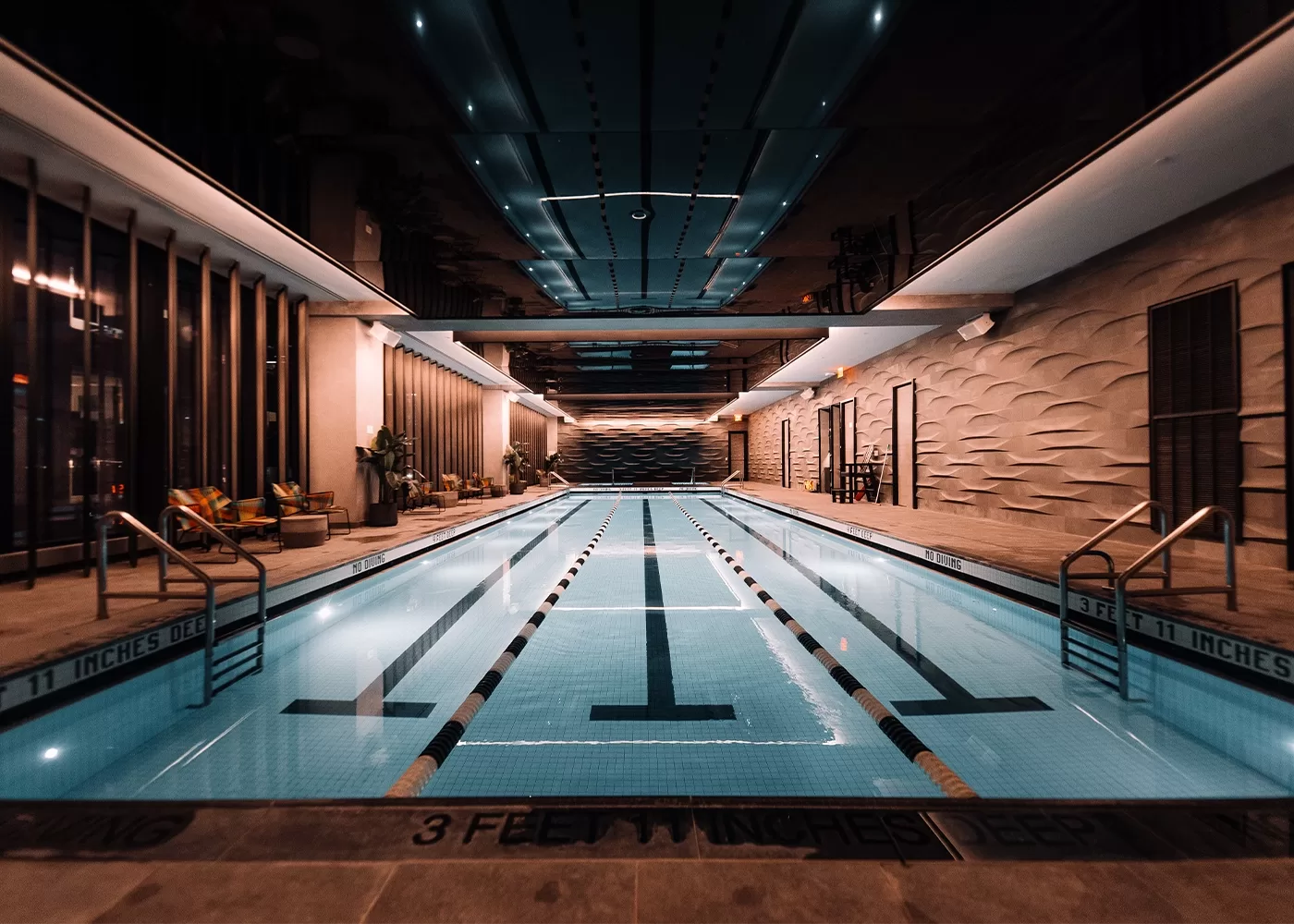 Large lit up swimming pool in a chrome coloured rom with a reflective ceiling showing the reflection of the pool. Floor to ceiling windows show New York in the dark.