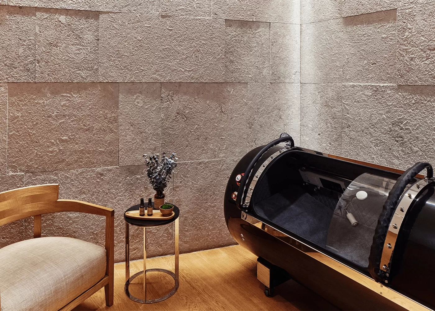 A minimalist room featuring a wooden chair with a beige cushion, a small round table with artful decorations, and a sleek black pod-like structure, all set against textured stone walls at Longevity at Bvlgari Spa.