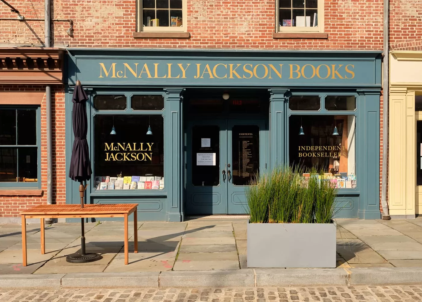 Exterior of McNally Jackson Books in NYC with a green facade and gold lettering. The storefront features large windows displaying books, a small table, a closed umbrella, and a planter with tall grasses on the cobblestone sidewalk.