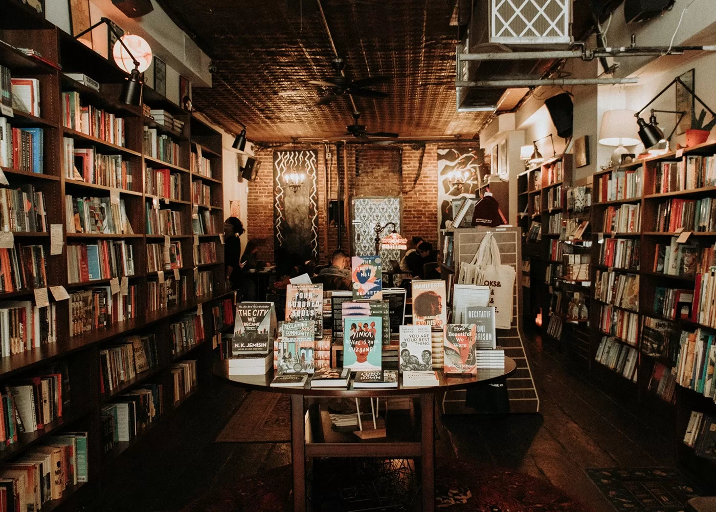 The cosy bookstore of Book Club Bar, East Village in NYC with, shelves filled with books on both sides. A central table displays various books. The lighting is warm, with a patterned wall and relaxed ambiance, suggesting a welcoming place for book lovers.