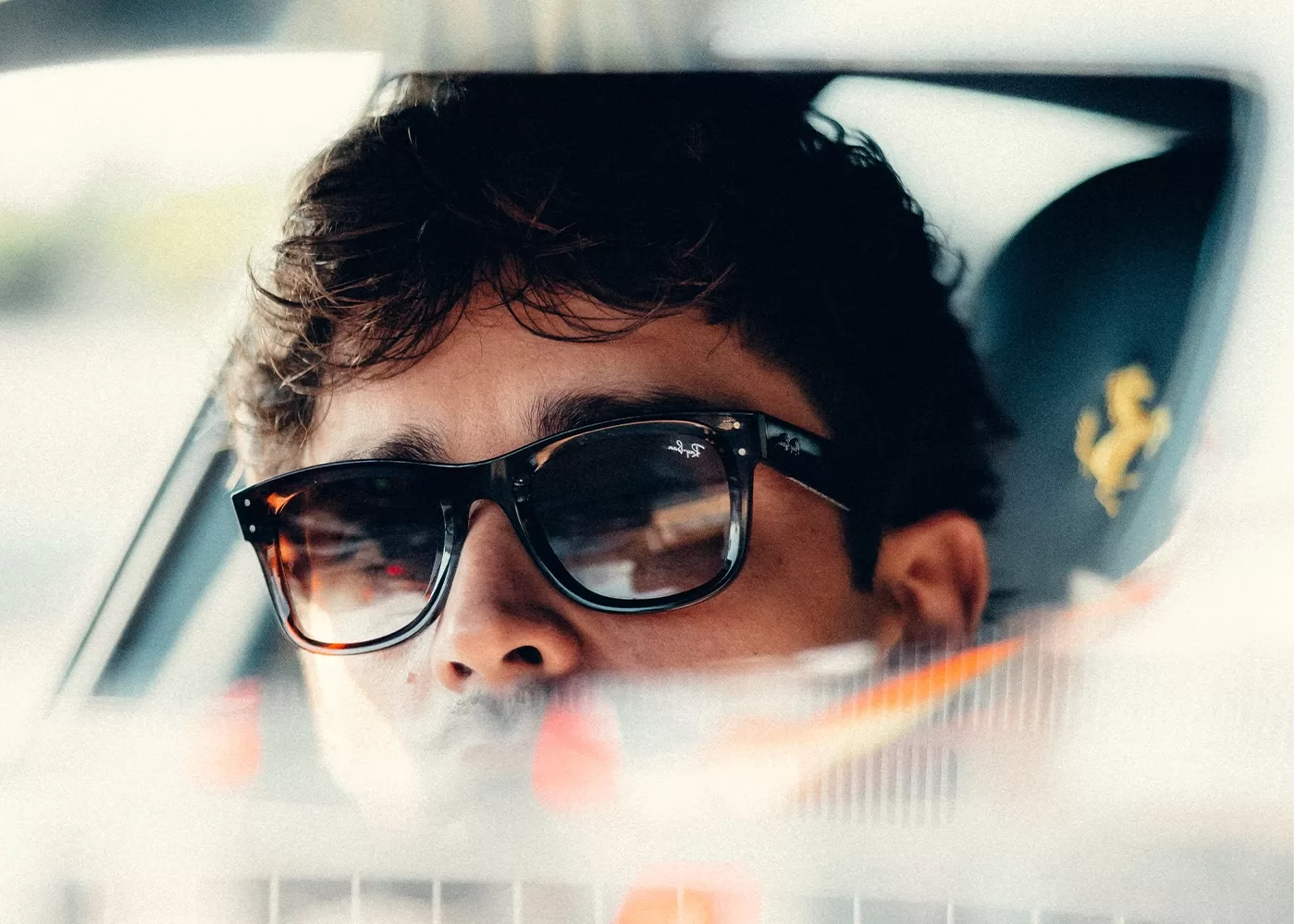 Close-up of Charles Leclerc wearing sunglasses, reflected in a cars rear-view mirror. His expression is relaxed, with soft lighting highlighting his face. A car seat with a logo is visible in the blurred background.