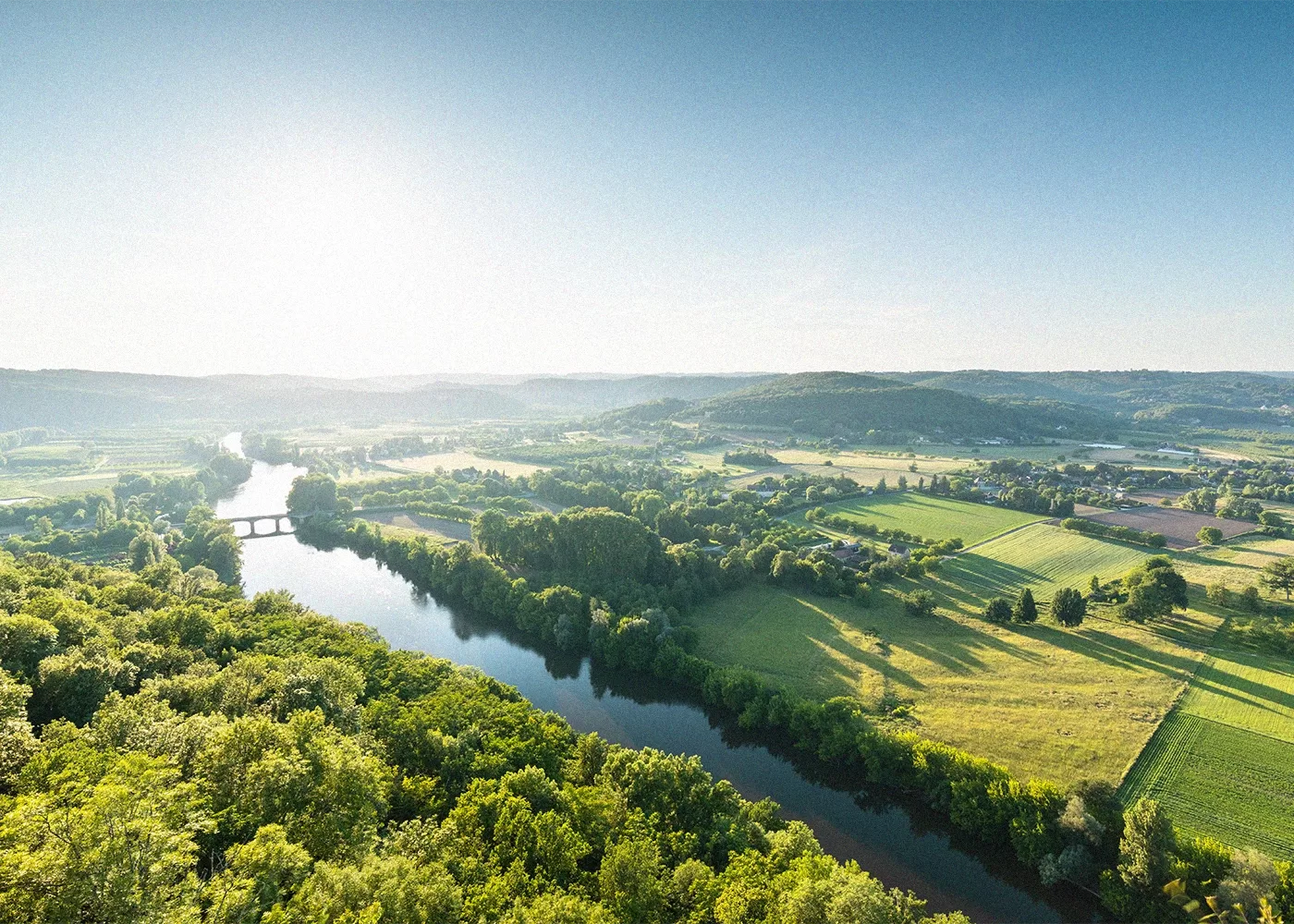 A scenic view of a river meandering through lush green fields and forests under a clear blue sky. A bridge spans the river, with rolling hills and farmland visible in the background in France.