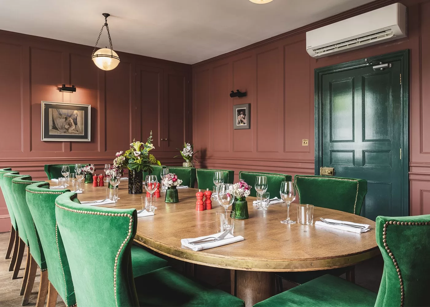 A dining room with a large oval wooden table, green velvet chairs, place settings with glasses and flowers, red paneled walls, framed art, and a dark green door. An air conditioner is mounted above the door.