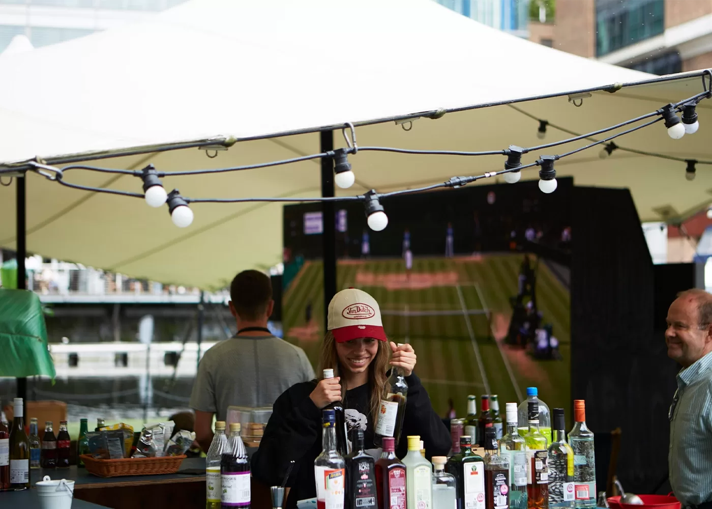 A smiling woman in a baseball cap stands behind an outdoor bar with various bottles. String lights hang above, and a tennis match is shown on a large screen in the background. Two men are also nearby.