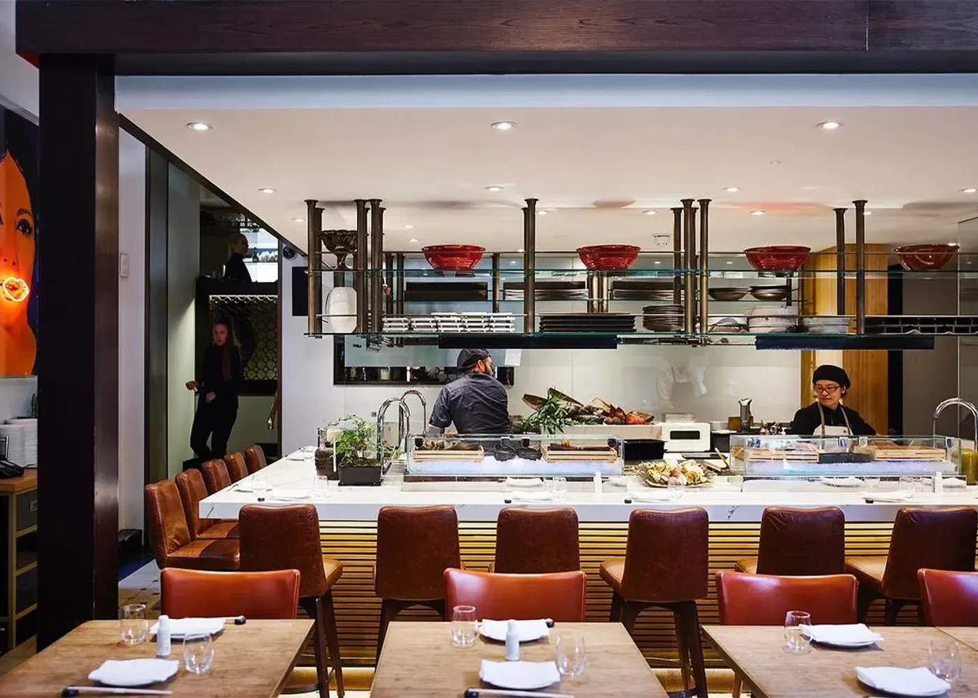 A modern restaurant interior with an open kitchen. Chefs are preparing food behind a white marble counter. Red bowls and plates are displayed on shelves above. The dining area features wooden tables and brown leather chairs at Dinings SW3, London.
