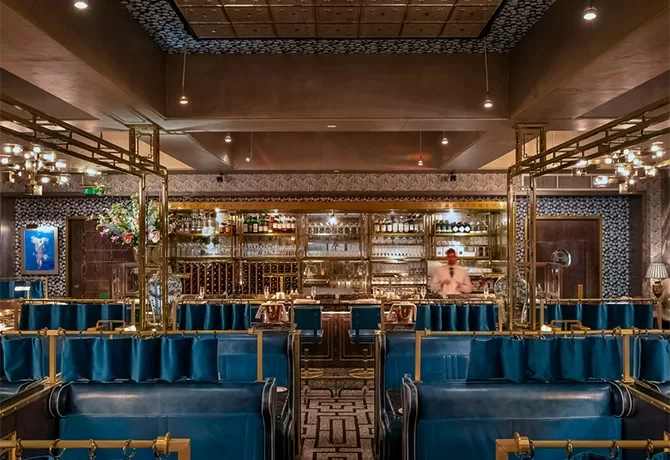 Luxurious restaurant interior with blue velvet seating, gold accent decor, and a patterned floor. A bartender stands behind a well-stocked, illuminated bar with glass shelves in the background at Bob Bob Ricard Soho London, London.