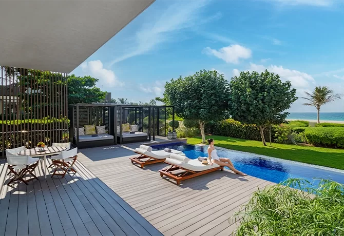 A woman relaxes on a lounge chair by an outdoor pool overlooking the ocean. The scene includes lush greenery, modern cabanas, and a small dining table on a wooden deck under a clear blue sky at The Oberoi Beach Resort, Al Zorah, Dubai.