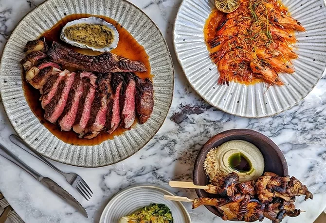 A marble table with plates of food: sliced steak with sauce, grilled prawns with lemon garnish, skewered grilled chicken with hummus, and a small bowl of greens. Silverware is placed beside the plates at Milk Beach, London.