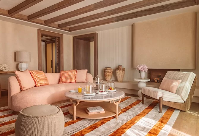 A cosy living room with a pink sofa and orange-checkered pillows. Theres a round coffee table with drinks and snacks, a striped armchair, and decorative vases. The room features wooden beams and a patterned rug in warm tones in Temuda Bay.
