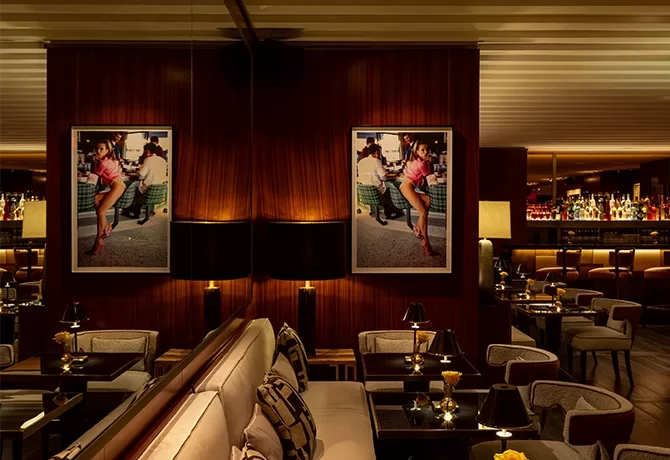 A dimly lit, upscale lounge interior with dark wood walls and cozy seating. A large photograph of a woman dancing is prominently displayed. Small lamps on tables create a warm ambiance, and a bar with various bottles is seen in the background.