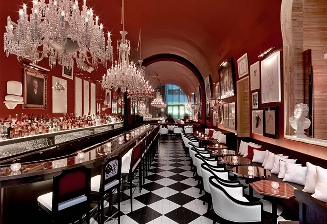 Elegant bar and restaurant with high, arched ceilings and red walls adorned with art. Crystal chandeliers hang above a long bar and black-and-white checkered floor. White cushioned chairs at tables create a sophisticated atmosphere.