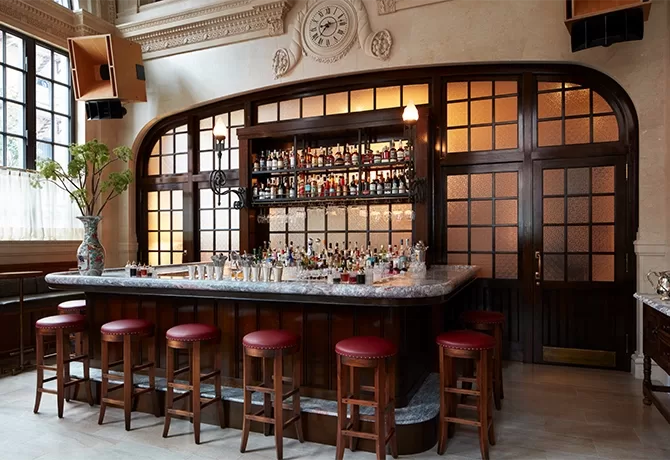 A stylish bar with a marble countertop, surrounded by red cushioned stools. Shelves behind display various bottles. The room features large windows, soft lighting, and elegant wood paneling with decorative accents on the walls and ceiling.