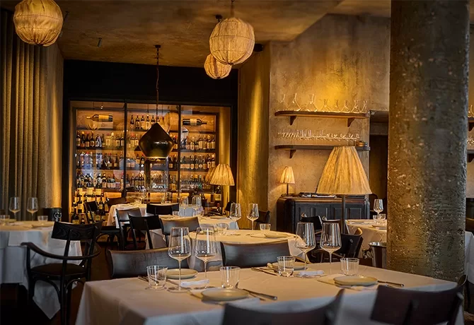 A dimly lit, elegant restaurant with white tablecloths, glassware, and dark chairs. Warm lighting from unique pendant lamps and a wall of shelves holding bottles creates an inviting ambiance at Fantômas, London.