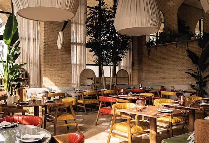 A cosy restaurant interior with rustic brick walls and large windows. There are wooden tables and colorful cushioned chairs, surrounded by plants. Large pendant lights hang from the high ceiling, creating a warm and inviting atmosphere at The Campaner, London.