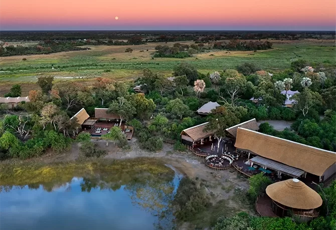 Aerial view of a safari lodge surrounded by lush greenery and a waterhole. Thatched-roof buildings are nestled among trees. The horizon shows a colorful sunset with a rising moon. Open plains stretch into the distance.