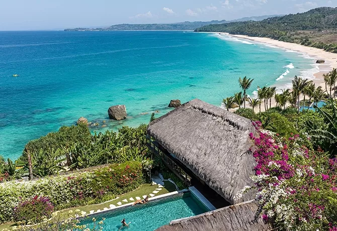A stunning coastal view features a thatched-roof villa with a swimming pool surrounded by lush greenery and colorful flowers. Beyond, a pristine white sand beach meets turquoise waters, with a backdrop of hills under a clear blue sky.