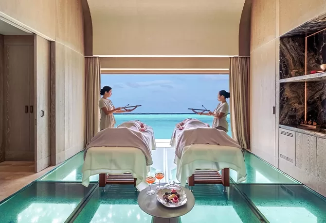 Two people receive spa treatments on massage tables in a luxurious room with glass floors. Two spa attendants stand beside them. A large window provides a view of the ocean, creating a serene atmosphere. A small table holds spa essentials.