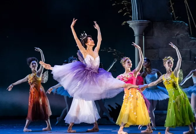 A ballerina in a purple and white tutu stands on stage, surrounded by dancers in tutus. They hold sparkling wands, poised elegantly on a staircase backdrop, creating a magical, fairytale-like scene at Royal Ballet & Opera’s Cinderella.
